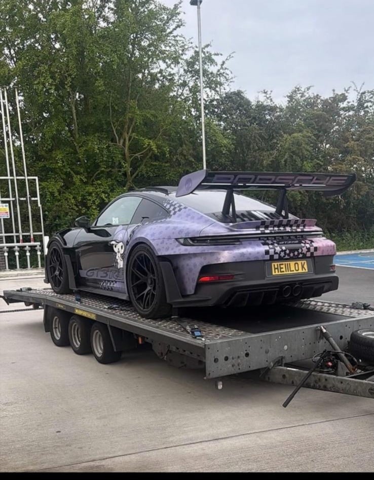 Purple Porsche 911 with wide body kit and large rear wing being transported on a flatbed trailer