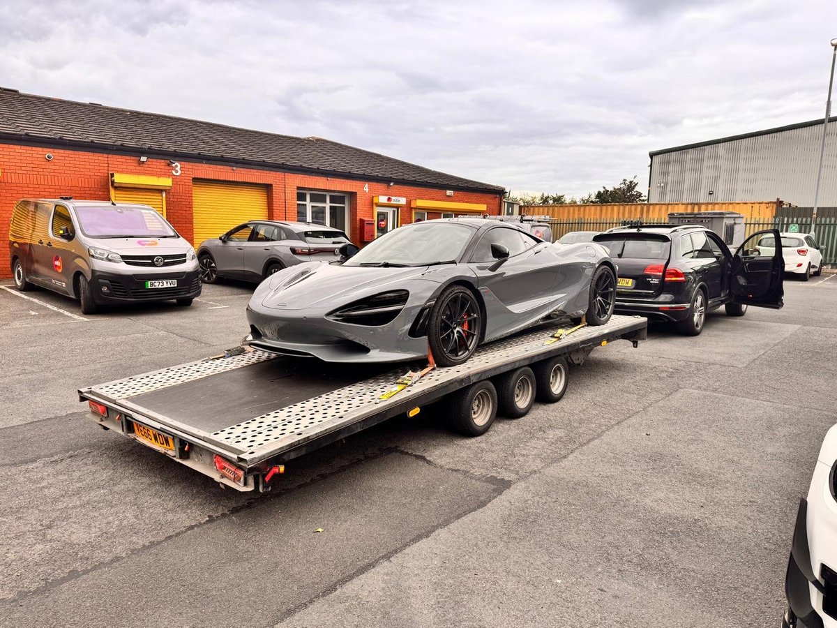 Gray McLaren 720S sports car on a flatbed trailer at a dealership with other vehicles and an orange building in background